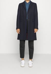 Marineblauwe wollen jas met een getailleerd ontwerp, voorzien van geknipte revers, twee voorzakken en een enkele knoopsluiting. Gecombineerd met een spijkerhemd en zwarte jeans.