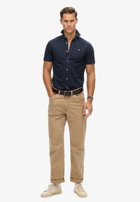 Marineblauw overhemd met korte mouwen en borstzak, gecombineerd met beige broeken en lichte sneakers. Heeft een eenvoudig, klassiek ontwerp.