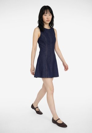 Mujer caminando hacia adelante con un vestido vaquero azul oscuro sin mangas y zapatos Mary Jane marrón oscuro sobre un fondo blanco.