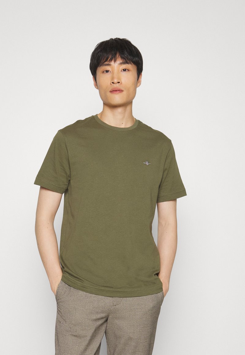 Tricou verde măsliniu, cu mânecă scurtă, dintr-un material moale, având un decolteu rotund și un logo mic pe piept. Modelul stă cu mâinile în buzunare.