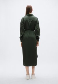 Μακρύ, σκούρο πράσινο φόρεμα με ζώνη στη μέση, γιακά και μακρυά μανίκια, κατασκευασμένο από λείο ύφασμα. Διαθέτει μήκος μέχρι τη μέση της γάμπας και πλευρική σκισμή.