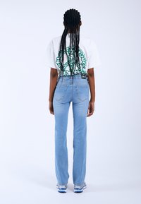 Jeans de mezclilla azul claro con un diseño de pierna recta, textura desgastada y cinco bolsillos, combinados con una camiseta blanca que presenta un detalle gráfico en verde.