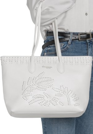 Sac cabas en cuir blanc avec motif de feuilles découpées, double anse épaule, porté par une personne en chemisier blanc et jean bleu avec ceinture noire.