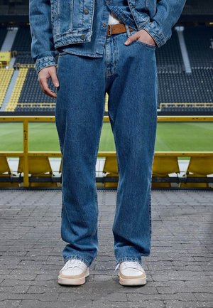 Man draagt een blauw spijkerjack en spijkerbroek met een bruine riem en witte sneakers, staand op een betegeld gebied vlak bij een tribune van een stadion.