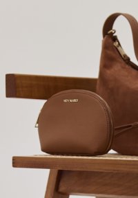 Een ronde bruine leren tas met een rits en gouden accenten ligt naast een bruine suède tas op een houten bank. Logo "HEY MARLY" zichtbaar.