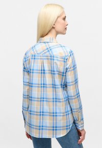 Frau mit langen blonden Haaren, die ein blaues und beiges kariertes Button-Up-Hemd und blaue Jeans trägt. Sie steht seitlich, mit sichtbarem Rücken.