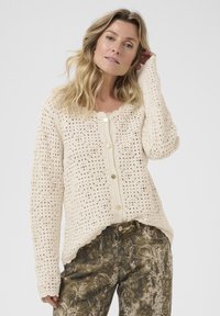 Blonde Frau, die eine cremefarbene gehäkelte Strickjacke mit goldenen Knöpfen und gemusterten dunkelgrünen und braunen Hosen trägt, und mit einer Hand nahe ihrem Kopf posiert.