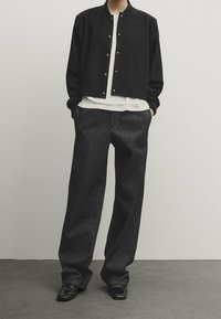 Personne portant une veste noire boutonnée, une chemise blanche, un jean sombre ample et des chaussures noires, debout les mains dans les poches contre un mur uni.