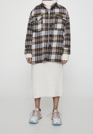 Person trägt eine grau-gelbe karierte, geknöpfte Jacke über einem weißen, gestrickten Rock und weiße Sneakers mit blauen Sohlen und mehrfarbigen Schnürsenkeln.