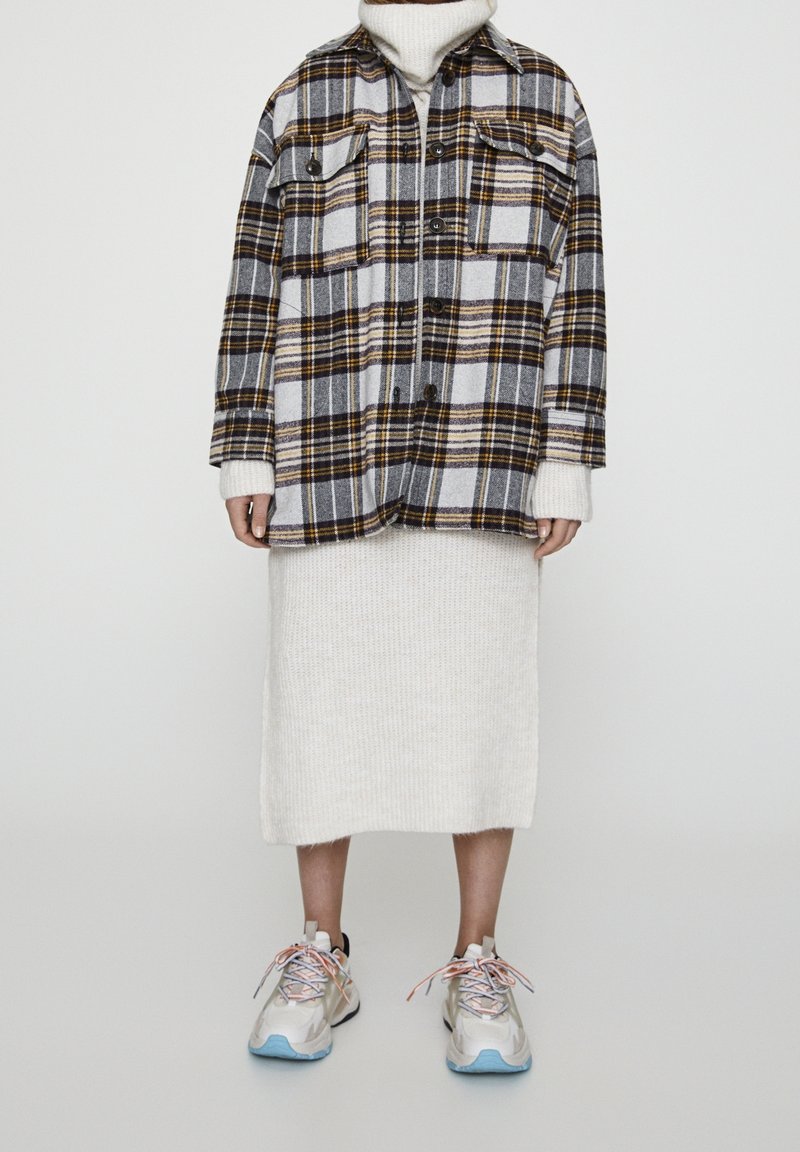 Personne portant une veste boutonnée à carreaux grise et jaune par-dessus une jupe blanche tricotée et des baskets blanches avec des semelles bleues et des lacets multicolores.