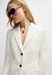 White tailored blazer with two black buttons, notched lapels, and side pockets. Worn with black sunglasses and a silver chain necklace featuring a red heart pendant.