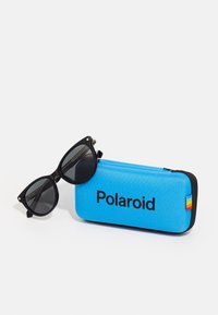 Occhiali da sole a forma di occhi di gatto neri accanto a una custodia blu con zip che presenta il logo "Polaroid" e un dettaglio a strisce multicolori.
