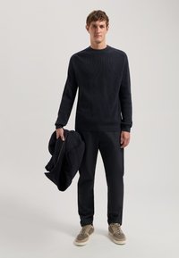 Pulover tricotat negru cu textură cu nervuri, asortat cu pantaloni negri. Bărbatul ține o jachetă închisă la culoare într-o mână și poartă adidași gri.