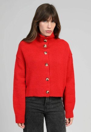 Femme portant un pull à col roulé rouge boutonné avec des manches longues et un pantalon noir sur fond gris uni.
