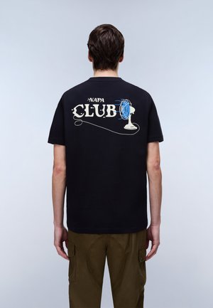 Moški, ki nosi črno majico s tekstom "NAPA CLUB" in modro grafiko električnega ventilatorja na hrbtni strani, v kombinaciji s rjavimi hlačami.