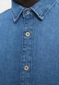 Világoskék farmering klasszikus gallérral, két gombbal a gombsoron, és varrási díszítéssel a varrások mentén.