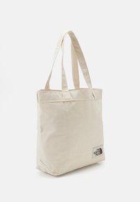 Bolso tote beige hecho de lona con dos asas de tela, que presenta un pequeño parche con logo en el lateral. Diseño simple con forma rectangular.