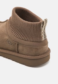 Botte de cheville en suede marron avec col en tricot côtelé, dotée d'une étiquette décorative et d'une semelle texturée. Logo embossé sur le côté.