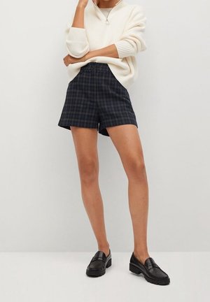 Shorts à carreaux noirs et bleu marine avec une coupe ajustée et une taille plate, associés à un pull en maille crème, et des mocassins noirs.