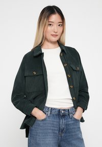 Mørkegrønn kordfløyelsjakke med krage, med brystlommer og messingknapper, brukt over en hvit skjorte og blå jeans.