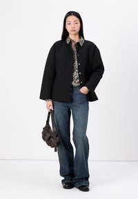 Must mustritud must jope nööpidega, lillemustriga särk ja laiad sinised teksad. Mudel hoiab väikest pruuni kotti; mustad kingadComplete the outfit.