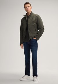 Giacca imbottita verde oliva con colletto alto, chiusura a zip e due tasche laterali, abbinata a jeans blu scuro e sneakers bianche.