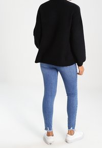 Persoon met een zwarte trui, lichtblauwe skinny jeans met rafelige zoom en witte sneakers, staand met de rug naar voren op een effen witte achtergrond.