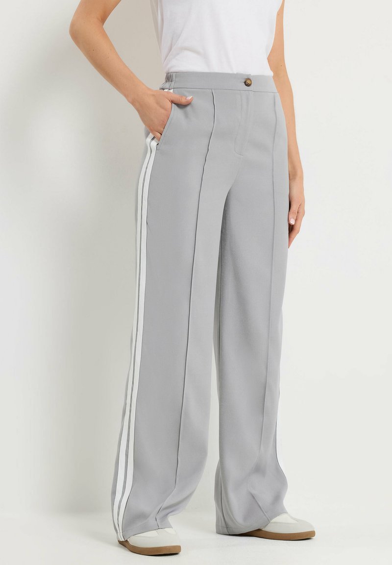 Person wearing light gray wide-leg pants with white side stripes, white shoes, and a white sleeveless top, standing with one hand in pocket.