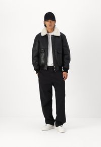 Černá kožená bomber bunda s ovčími límcem, spárovaná s bílým tričkem, černými kalhotami a bílými teniskami. Minimalistický design.