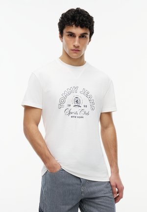 Ung mand iført en hvid "Tommy Jeans Sports Club New York" t-shirt og blå og hvide stribede bukser, stående med den ene hånd i lommen.