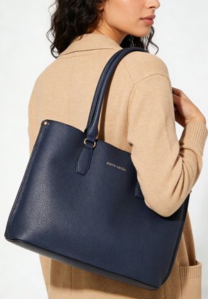 Vrouw in beige jas draagt een grote marineblauwe leren tas met dubbele handvatten op haar schouder.
