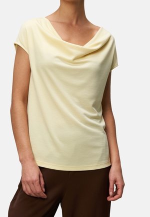 Vrouw in lichtgele korte mouw top met gedrapeerde halslijn en bruine broek, poserend tegen een effen lichte achtergrond.