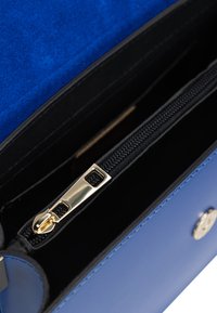 Modrá semišová clutch kabelka se zlatými detaily, obsahující zipovou černou kapsu a hladký povrch s kontrastním prošitím.