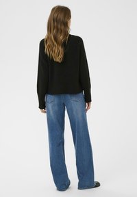 Kvinde med langt bølget hår iført en sort sweater, blå brede bukser og sorte sko, stående med ryggen til på en ensfarvet baggrund.