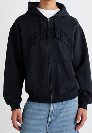 Mann trägt schwarzen Kapuzenpullover mit Reißverschluss und dem Schriftzug "GAP ATHLETIC", kombiniert mit hellblauen Jeans.