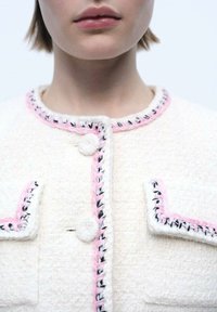 Witte tweedjas met contrasterende roze en zwarte stiksels, voorzien van twee voorzakken en ronde, gestructureerde knopen.