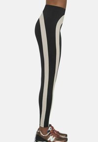 Legginsy widok z boku, wysoki stan, czarna tkanina i szerokie pionowe beżowe paski, noszone z brązowymi trampkami New Balance.