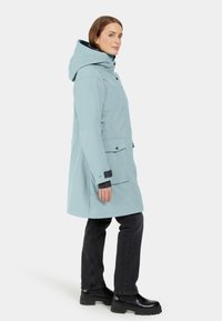 Ljusblå vattentät parka med huva, sidofickor och svarta ärmaccent. Buren över mörka byxor och kraftiga svarta stövlar.