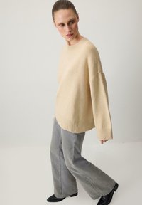 Béžový pletený sveter s voľným strihom, rebrovaným lemom a spadlými ramenami, kombinovaný so svetlošedými rozšírenými džínsami a čiernymi členkovými topánkami.