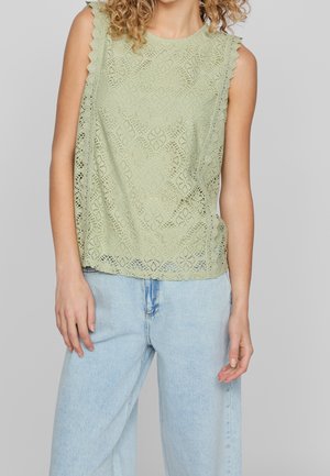 Haut sans manches vert clair à motif de dentelle avec bords festonnés, associé à un jean en denim bleu clair. Tissu texturé avec une coupe ample.