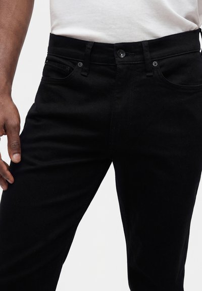 Gros plan d'une personne portant un jean noir et une chemise blanche, montrant la zone avant de la taille et une partie de la main gauche.