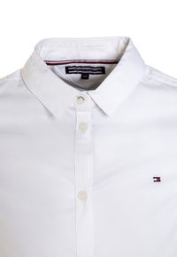 Weiße Bluse mit Knopfleiste und kleinem gesticktem Logo mit rot, weiß und blauen Akzenten auf der linken Brust.