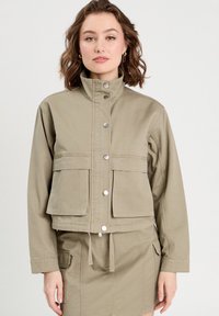 Chaqueta corta color caqui hecha de tela de algodón con un cuello alto, botones de presión y dos bolsillos frontales. Cuerda ajustable en el dobladillo inferior.