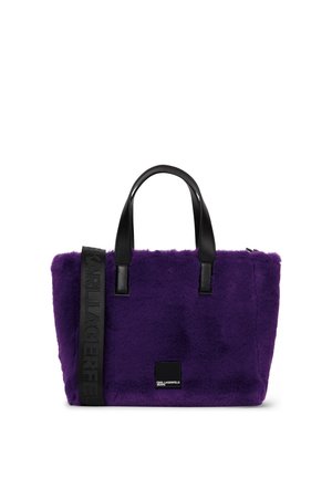 Sac cabas en fausse fourrure violette avec poignées noires et une sangle noire embossée du nom de la marque, avec un petit patch logo noir à l'avant.