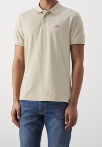 Béžové polo tričko s límečkem, třemi knoflíky a malým červeným logem Levi's na hrudi. Nošeno s modrými denimovými džíny.