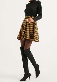 Mujer con una blusa negra de manga larga, falda de pata de gallo amarilla y negra, medias negras transparentes y botas de tacón altas negras hasta la rodilla.