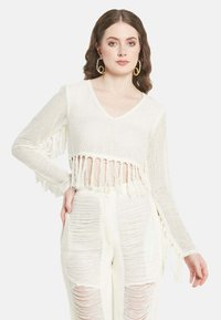 Weißer, gekürzter Pullover mit V-Ausschnitt, Quastenverzierungen am Saum und den Ärmeln, kombiniert mit strukturierten, weißen Hochbundhosen.
