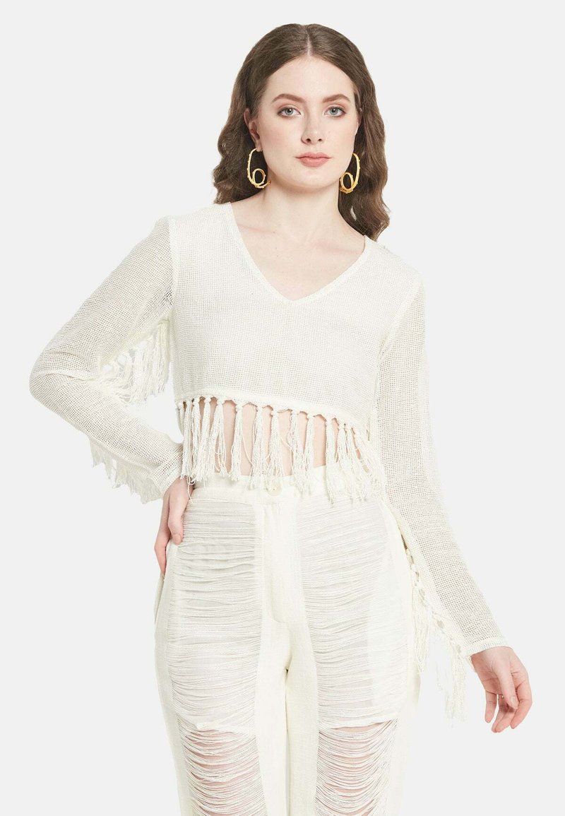 Weißer, gekürzter Pullover mit V-Ausschnitt, Quastenverzierungen am Saum und den Ärmeln, kombiniert mit strukturierten, weißen Hochbundhosen.