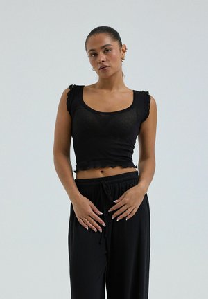 Kvinde med trukket hår tilbage, iført en sort ærmeløs crop top med flæser og sorte bukser med snøre, stående mod en ensfarvet baggrund.