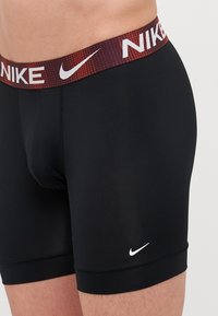 Črne športne kratke hlače s prilegajočo zasnovo in rdečim pasom z vzorcem, na katerem je napis "NIKE" v beli barvi. Vključujejo tudi majhen bel logo na nogi.